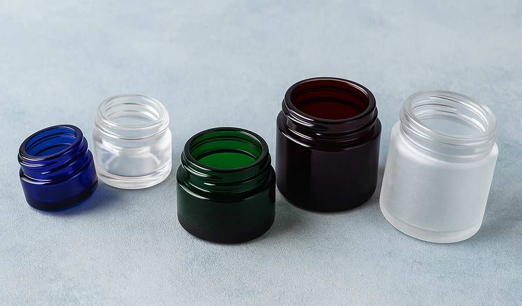 Assorted open glass cosmetic jars in blue, clear, green, amber, and frosted finishes arranged on a soft blue textured background.